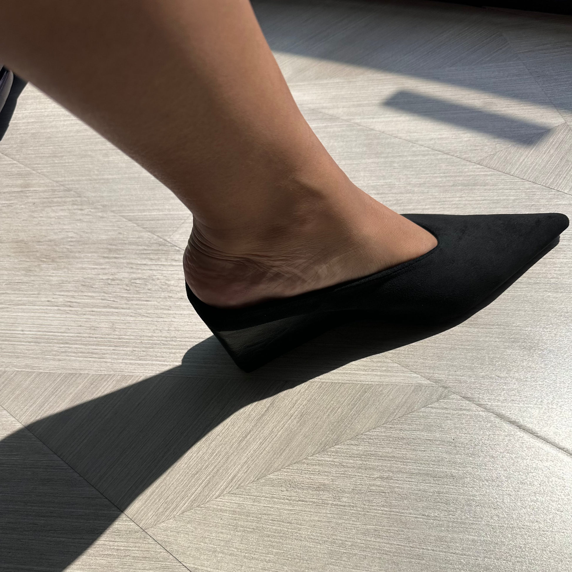 Person wearing a black suede mule pointed sandal on a light wooden floor